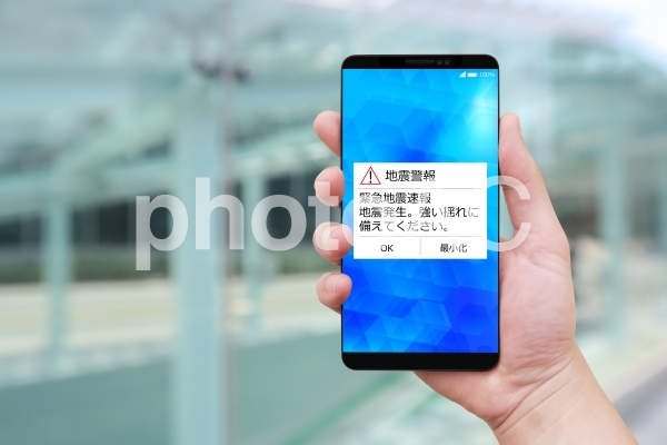 緊急地震速報を受信するスマホを持つ男性の手-街並み後ろボケ背景 緊急地震速報を受信するスマホを持つ男性の手-街並み後ろボケ背景の写真